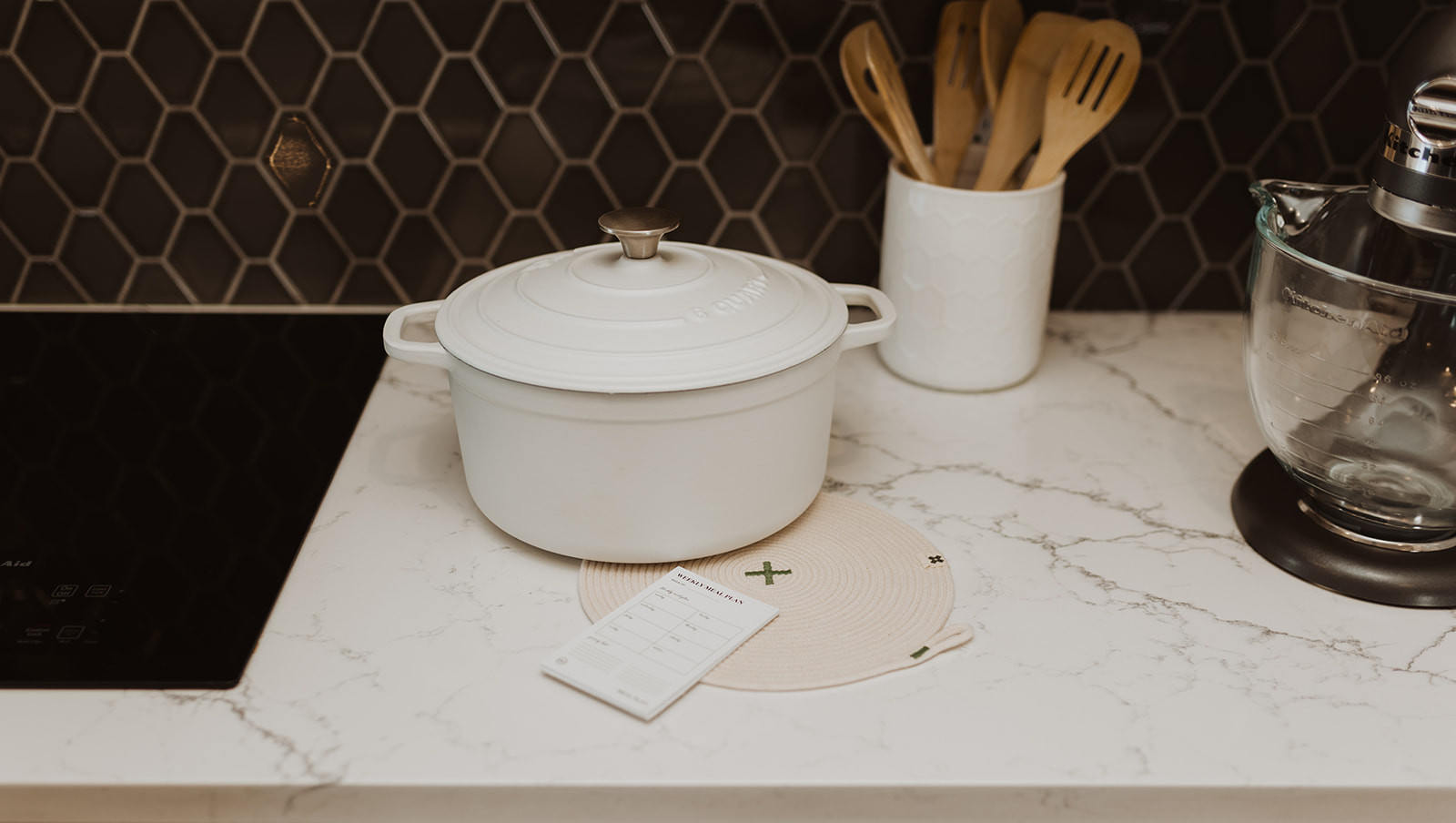 kitchen counter with white pot, wooden utensils and black tiled backsplash